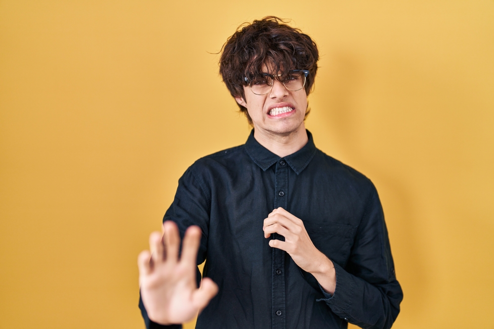 Young man grossed out with hands in front of him looking at the camera in front of yellow background