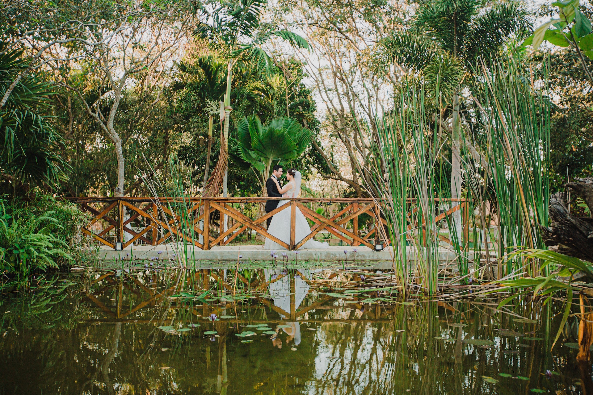 bride and groom standing on a bridge next to a pond